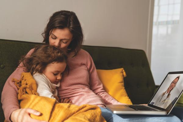 Moeder troost haar jonge dochter terwijl ze samen op de bank zitten, met een laptop waarop een arts op het scherm te zien is.